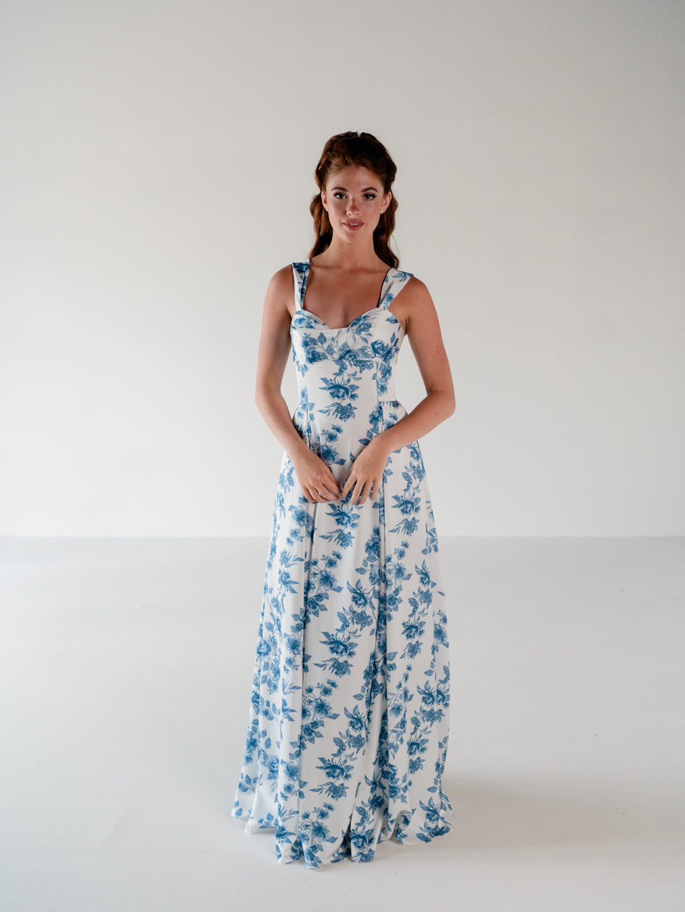 A woman modeling a sleeveless white maxi dress with a blue floral toile pattern, standing against a plain white studio background. / Draped neckline A-line gown with faux overskirt and slit detail.