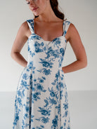 A woman wearing a sleeveless white dress with a blue floral toile pattern, photographed against a plain light gray studio background. / Draped neckline A-line gown with faux overskirt and slit detail.