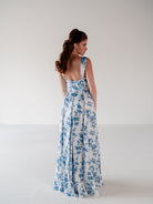 A woman wearing a sleeveless white maxi dress with a blue floral print, shown from the back in a studio setting with a plain light gray background. / Draped neckline A-line gown with faux overskirt and slit detail.