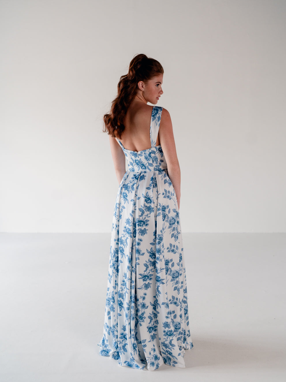 A woman wearing a sleeveless white maxi dress with a blue floral print, shown from the back in a studio setting with a plain light gray background. / Draped neckline A-line gown with faux overskirt and slit detail.