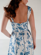 A woman shown from the back wearing a sleeveless white dress with a blue floral toile pattern and a low open back. / Draped neckline A-line gown with faux overskirt and slit detail.