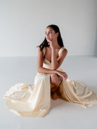 A woman kneels on a white studio floor wearing a flowing champagne satin dress with draped fabric pooling around her. / Draped neckline A-line gown with faux overskirt and slit detail.