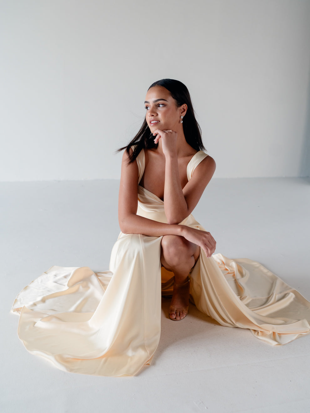 A woman kneels on a white studio floor wearing a flowing champagne satin dress with draped fabric pooling around her. / Draped neckline A-line gown with faux overskirt and slit detail.