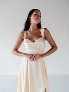 A woman modeling a sleeveless cream satin dress with a sweetheart neckline and draped bodice, set against a plain light gray studio background. / Draped neckline A-line gown with faux overskirt and slit detail.
