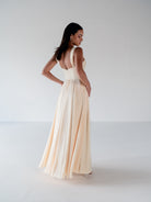 A woman modeling a floor-length cream or ivory formal gown from the back, standing in a bright, minimal studio with a white background. / Draped neckline A-line gown with faux overskirt and slit detail.