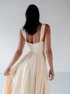 A woman shown from behind wearing a sleeveless cream-colored maxi dress with a deep open back and pleated skirt, set against a plain light gray studio background. / Draped neckline A-line gown with faux overskirt and slit detail.