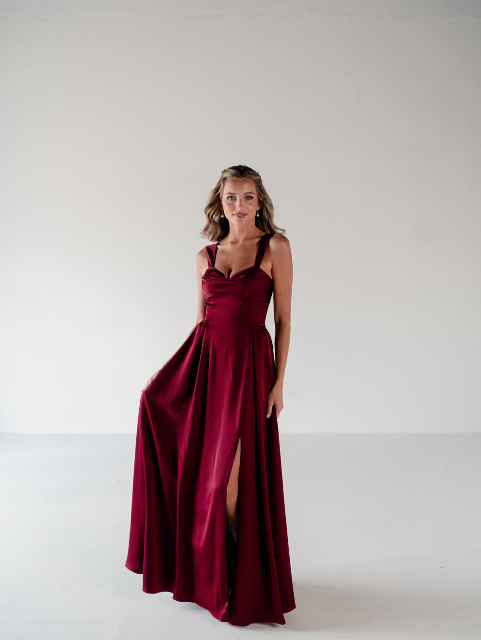 A woman modeling a floor-length burgundy satin gown with a draped neckline, halter-style straps, and a high thigh slit, standing against a plain light gray studio background. / Draped neckline A-line gown with faux overskirt and slit detail.