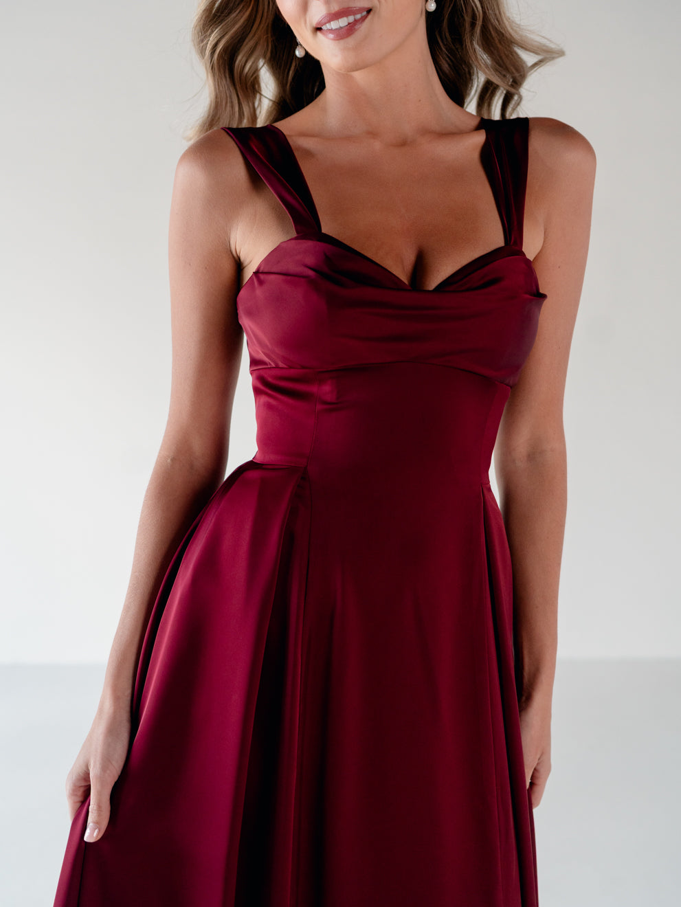A woman wearing a sleeveless, deep burgundy A-line dress with a draped sweetheart neckline and wide shoulder straps, photographed against a light gray studio background. / Draped neckline A-line gown with faux overskirt and slit detail.