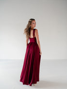 A woman modeling a floor-length burgundy satin gown from the back, standing against a seamless light gray studio background. / Draped neckline A-line gown with faux overskirt and slit detail.