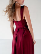 A woman wearing a sleeveless deep burgundy maxi dress with a structured open back and pleated skirt, shown from a rear three-quarter angle against a plain light background. / Draped neckline A-line gown with faux overskirt and slit detail.