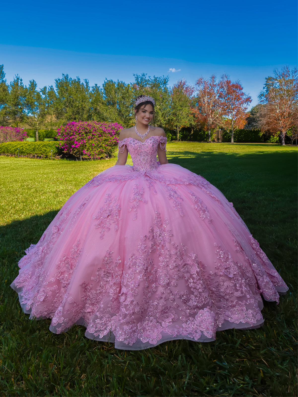 Woman in a pink ball gown standing outdoors with trees and grass in the background / This ball gown features a richly detailed bodice with floral appliqué and off-the-shoulder straps. The skirt is extremely full and layered with cascading floral accents, creating a dramatic, fairytale look.
