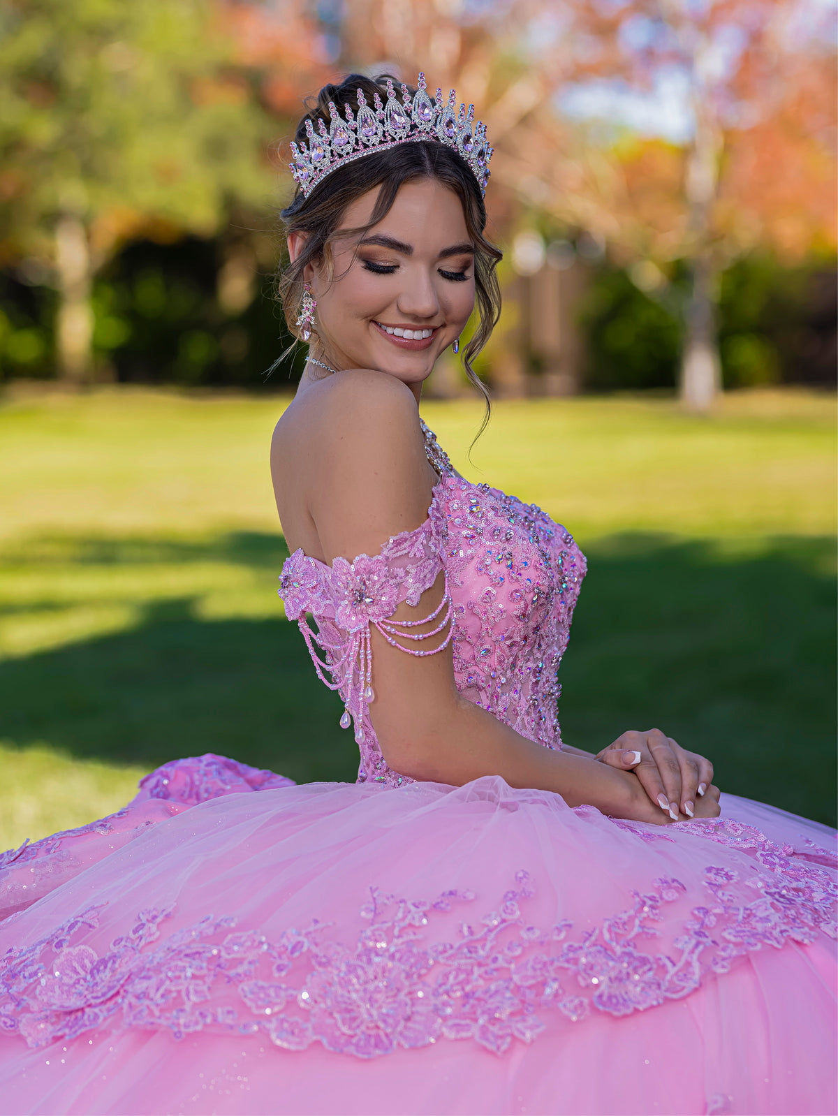 Woman in a pink quinceañera dress with a tiara outdoors. / This ball gown features a richly detailed bodice with floral appliqué and off-the-shoulder straps. The skirt is extremely full and layered with cascading floral accents, creating a dramatic, fairytale look.