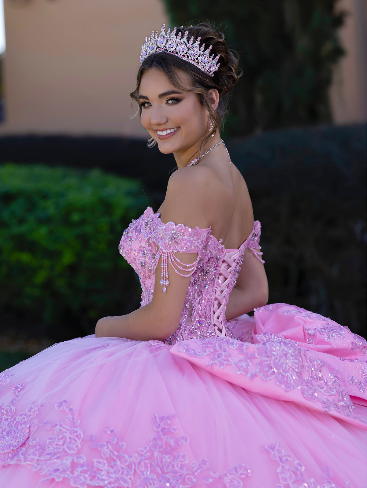 Woman in a pink quinceañera dress with a tiara, sitting outdoors. / This ball gown features a richly detailed bodice with floral appliqué and off-the-shoulder straps. The skirt is extremely full and layered with cascading floral accents, creating a dramatic, fairytale look.