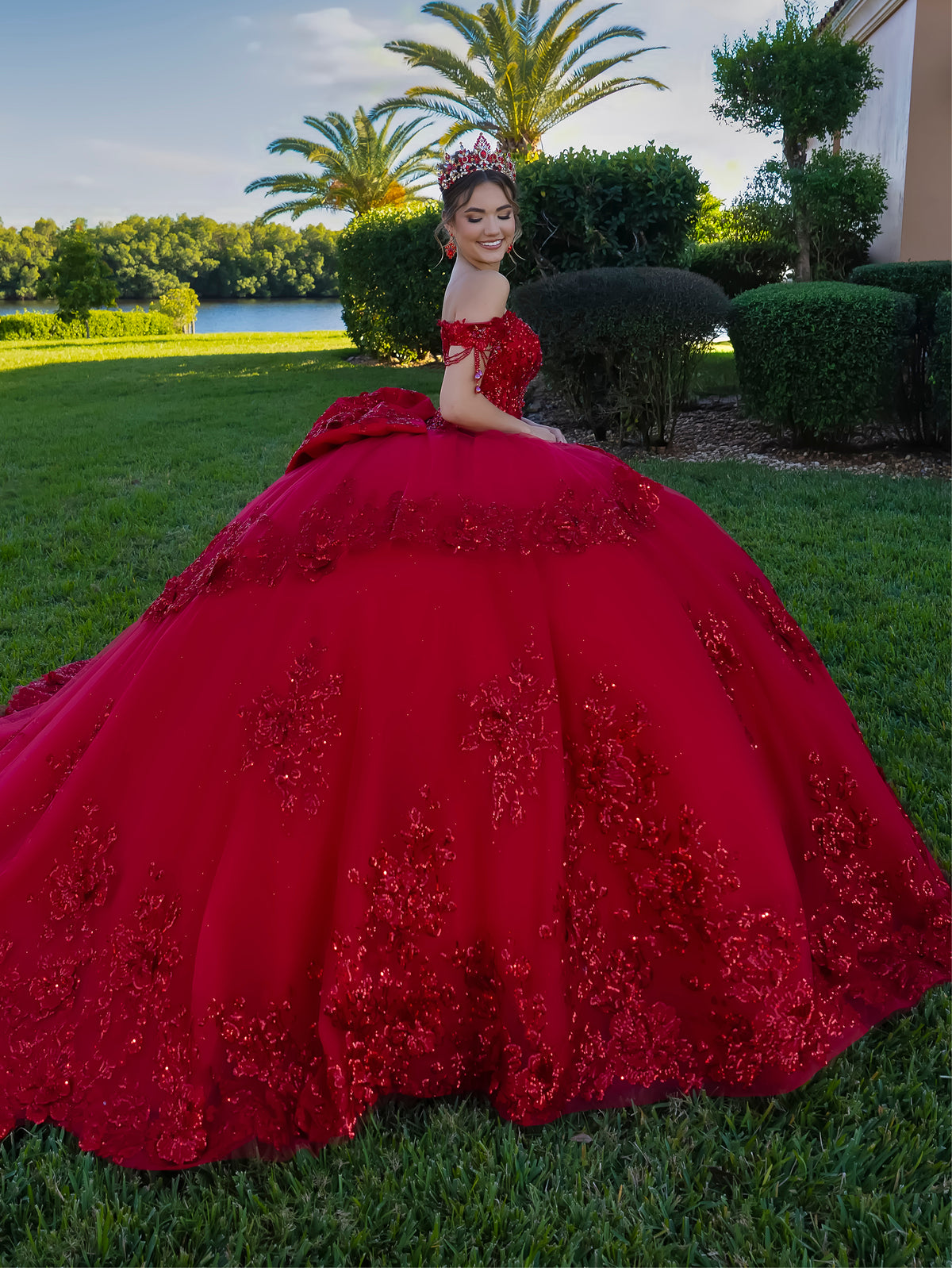 A young woman wearing a voluminous red ball gown with floral lace appliqué sits on a green lawn in front of palm trees and a lake. / This ball gown features a richly detailed bodice with floral appliqué and off-the-shoulder straps. The skirt is extremely full and layered with cascading floral accents, creating a dramatic, fairytale look.