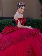 Woman in a red quinceañera dress with floral details and a tiara, sitting outdoors. / This ball gown features a richly detailed bodice with floral appliqué and off-the-shoulder straps. The skirt is extremely full and layered with cascading floral accents, creating a dramatic, fairytale look.
