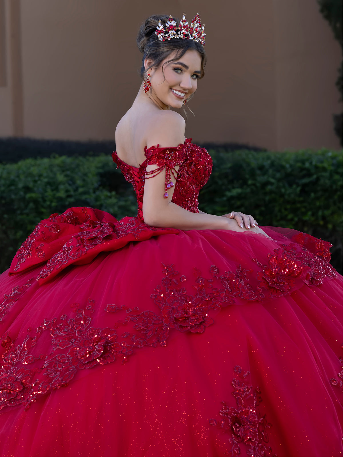 Woman in a red quinceañera dress with floral details and a tiara, sitting outdoors. / This ball gown features a richly detailed bodice with floral appliqué and off-the-shoulder straps. The skirt is extremely full and layered with cascading floral accents, creating a dramatic, fairytale look.