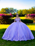 Woman in a purple ball gown standing in a garden with colorful flowers and greenery. / This ball gown features an ornate bodice with floral appliqué and off-the-shoulder straps. The skirt is extremely full of layered tulle and cascading floral details, creating a dramatic, fairytale silhouette.