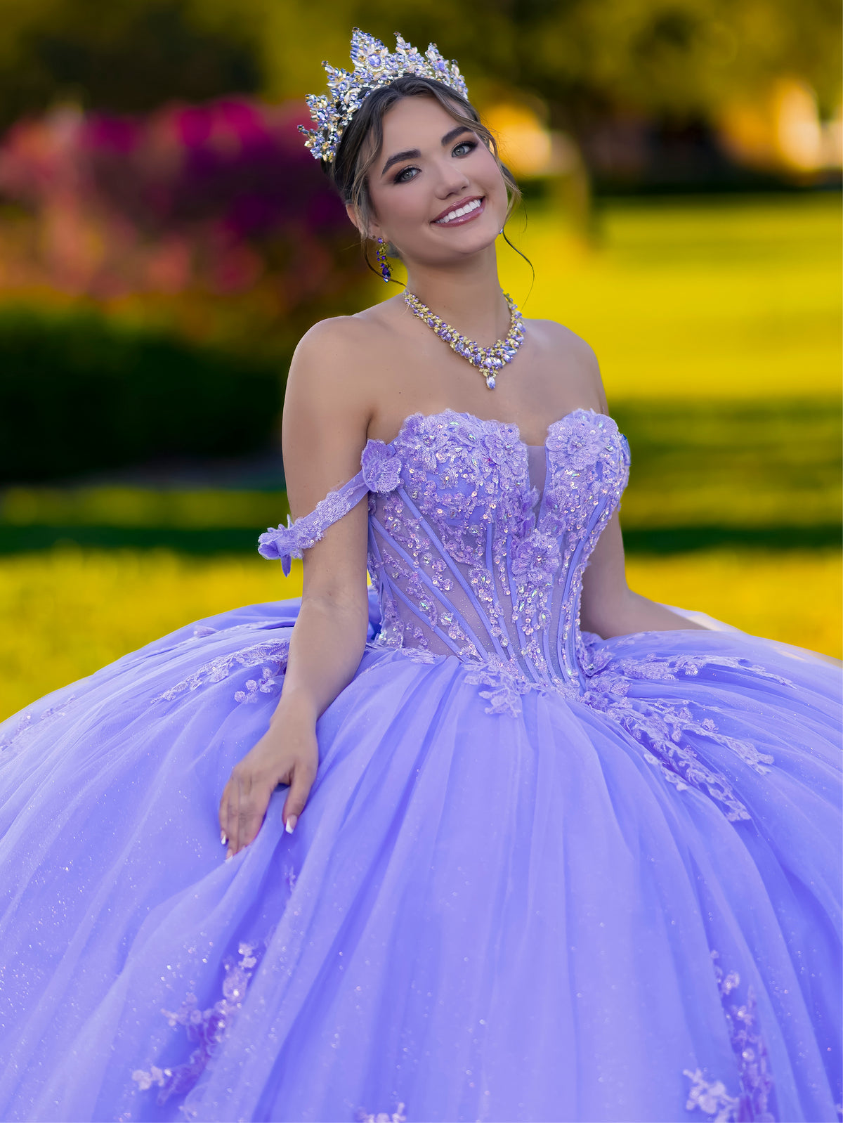 A young woman wearing a lavender off-the-shoulder quinceañera dress with a corset bodice, embellished tulle skirt, matching tiara, and necklace, posing outdoors in a sunny garden setting. / This ball gown features an ornate bodice with floral appliqué and off-the-shoulder straps. The skirt is extremely full of layered tulle and cascading floral details, creating a dramatic, fairytale silhouette.