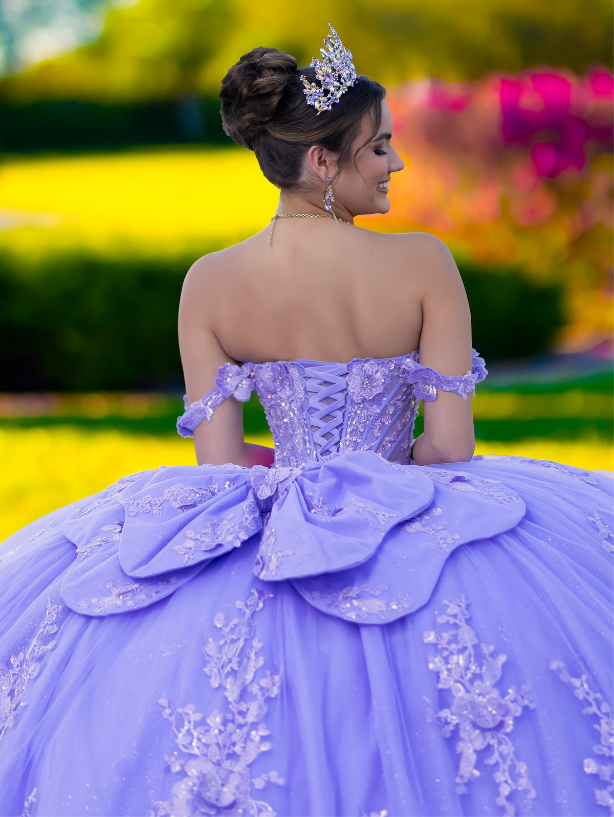 Woman in a purple quinceañera dress with floral details and a tiara, standing outdoors with colorful flowers in the background. / This ball gown features an ornate bodice with floral appliqué and off-the-shoulder straps. The skirt is extremely full of layered tulle and cascading floral details, creating a dramatic, fairytale silhouette.