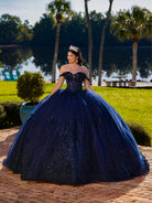 A woman wearing a voluminous off-the-shoulder navy blue ball gown with sparkling embellishments, a tiara, and a necklace, standing outdoors on a brick patio with a lake, palm trees, and green lawn in the background. / This ball gown features an ornate bodice with floral appliqué and off-the-shoulder straps. The skirt is extremely full of layered tulle and cascading floral details, creating a dramatic, fairytale silhouette.