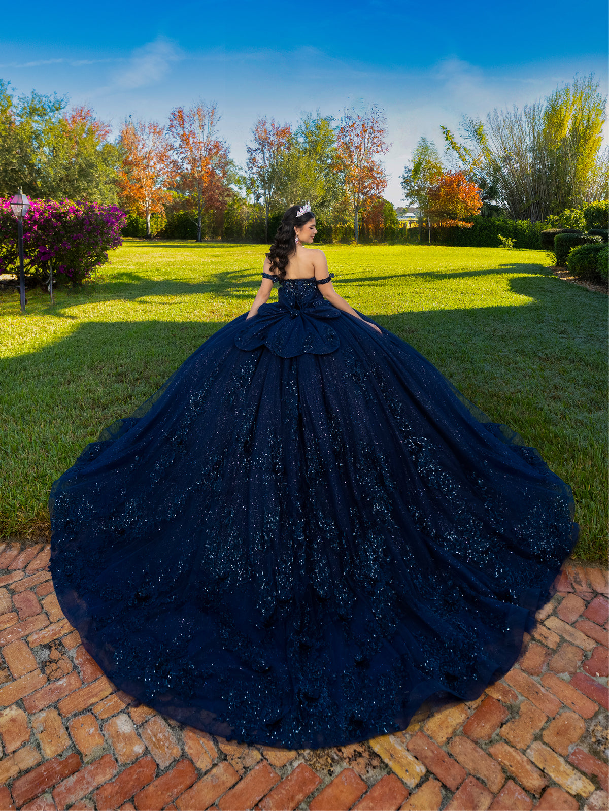 A woman stands outdoors on a brick path in a grassy park, wearing an elaborate off-the-shoulder ball gown with a very full dark navy skirt and train. Trees with green, orange, and red foliage are visible in the background under a blue sky. / This ball gown features an ornate bodice with floral appliqué and off-the-shoulder straps. The skirt is extremely full of layered tulle and cascading floral details, creating a dramatic, fairytale silhouette.