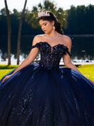 Woman in a navy blue sequin dress with a tiara outdoors / This ball gown features an ornate bodice with floral appliqué and off-the-shoulder straps. The skirt is extremely full of layered tulle and cascading floral details, creating a dramatic, fairytale silhouette.