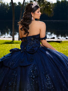A woman wearing a strapless navy blue quinceañera dress with sequined details and a large bow at the back, photographed outdoors near a lake with trees in the background. / This ball gown features an ornate bodice with floral appliqué and off-the-shoulder straps. The skirt is extremely full of layered tulle and cascading floral details, creating a dramatic, fairytale silhouette.