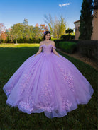 Woman in a purple floral dress standing outdoors with trees and a building in the background / This ball gown features a detailed floral-appliqué bodice with soft off-the-shoulder straps. The full layered skirt is adorned with matching floral designs and flows into a dramatic, elegant train.