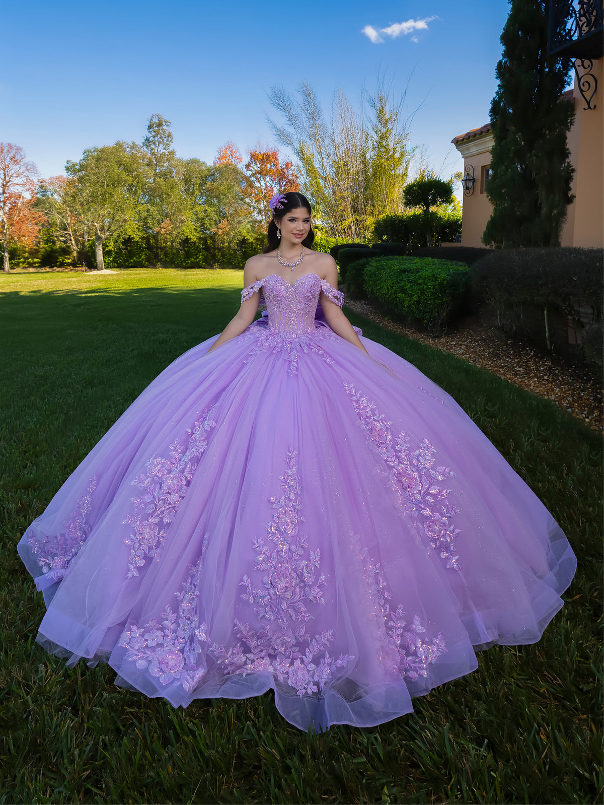 Woman in a purple floral dress standing outdoors with trees and a building in the background / This ball gown features a detailed floral-appliqué bodice with soft off-the-shoulder straps. The full layered skirt is adorned with matching floral designs and flows into a dramatic, elegant train.