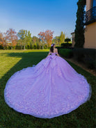 Person in a purple wedding dress standing on grass with trees and a building in the background / This ball gown features a detailed floral-appliqué bodice with soft off-the-shoulder straps. The full layered skirt is adorned with matching floral designs and flows into a dramatic, elegant train.