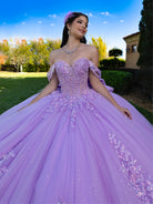 Woman in a purple ball gown with floral hairpiece outdoors. / This ball gown features a detailed floral-appliqué bodice with soft off-the-shoulder straps. The full layered skirt is adorned with matching floral designs and flows into a dramatic, elegant train.