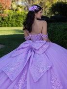 A woman photographed from behind wearing a lavender off-the-shoulder quinceañera dress with a large bow at the back, floral hair accessory, and sparkling embellishments, standing outdoors in a garden setting. / This ball gown features a detailed floral-appliqué bodice with soft off-the-shoulder straps. The full layered skirt is adorned with matching floral designs and flows into a dramatic, elegant train.