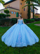 A woman wearing a voluminous light blue quinceañera ball gown stands on a green lawn in front of a stucco house with palm trees. The dress has off-the-shoulder straps, a fitted embellished bodice, and a very full tulle skirt with floral embroidery. / This ball gown features a detailed floral-appliqué bodice with soft off-the-shoulder straps. The full layered skirt is adorned with matching floral designs and flows into a dramatic, elegant train.