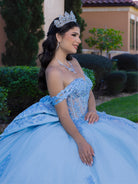A woman wearing a voluminous light blue quinceañera ball gown with off-the-shoulder straps, lace applique, and a sweetheart neckline, seated outdoors in front of trimmed hedges and a building. / This ball gown features a detailed floral-appliqué bodice with soft off-the-shoulder straps. The full layered skirt is adorned with matching floral designs and flows into a dramatic, elegant train.