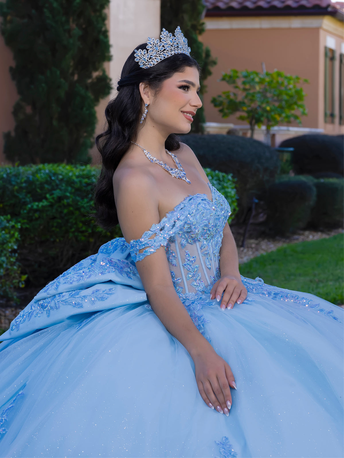 A woman wearing a voluminous light blue quinceañera ball gown with off-the-shoulder straps, lace applique, and a sweetheart neckline, seated outdoors in front of trimmed hedges and a building. / This ball gown features a detailed floral-appliqué bodice with soft off-the-shoulder straps. The full layered skirt is adorned with matching floral designs and flows into a dramatic, elegant train.