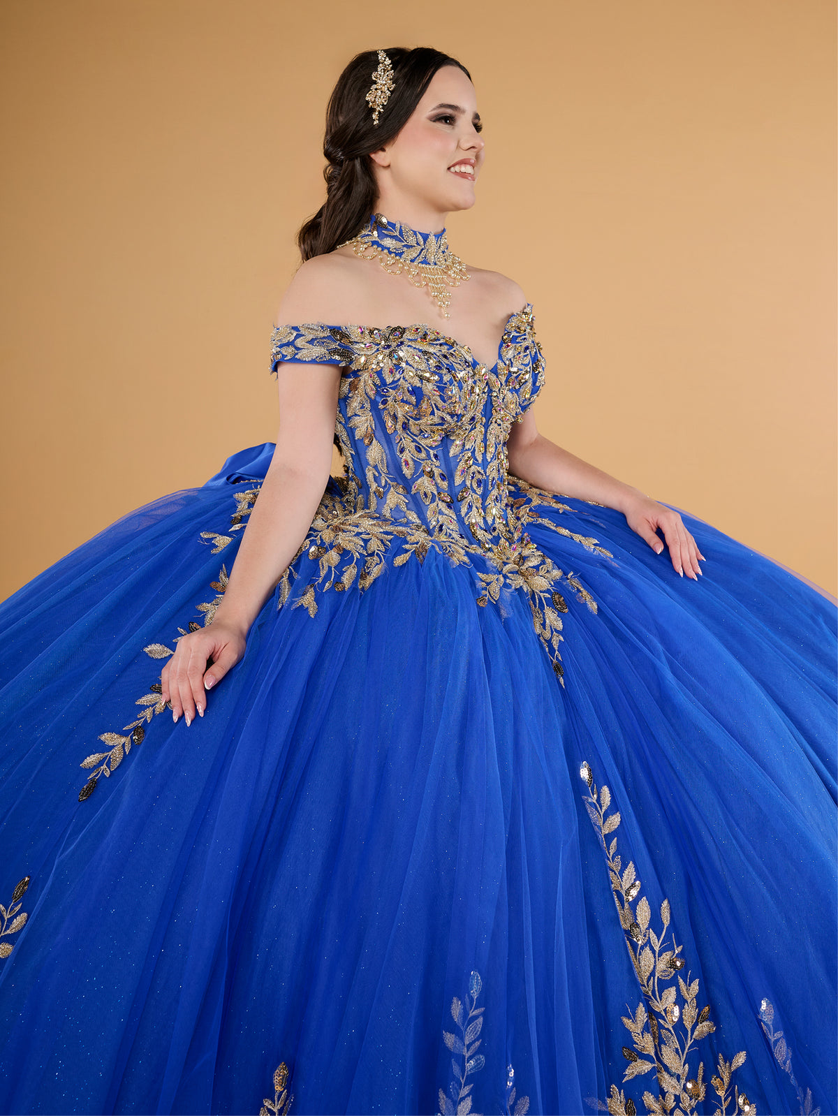 This ball gown features an ornate bodice with detailed embroidery and off-the-shoulder straps. The full tulle skirt is embellished with matching floral designs and finished with a dramatic bow and sweeping train.