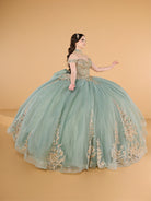 This ball gown features an ornate bodice with detailed embroidery and off-the-shoulder straps. The full tulle skirt is embellished with matching floral designs and finished with a dramatic bow and sweeping train.