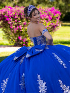 This ball gown features a detailed embroidered bodice and off-the-shoulder straps. The full, dramatic skirt is adorned with shimmering floral patterns and finishes with a large bow and flowing train.