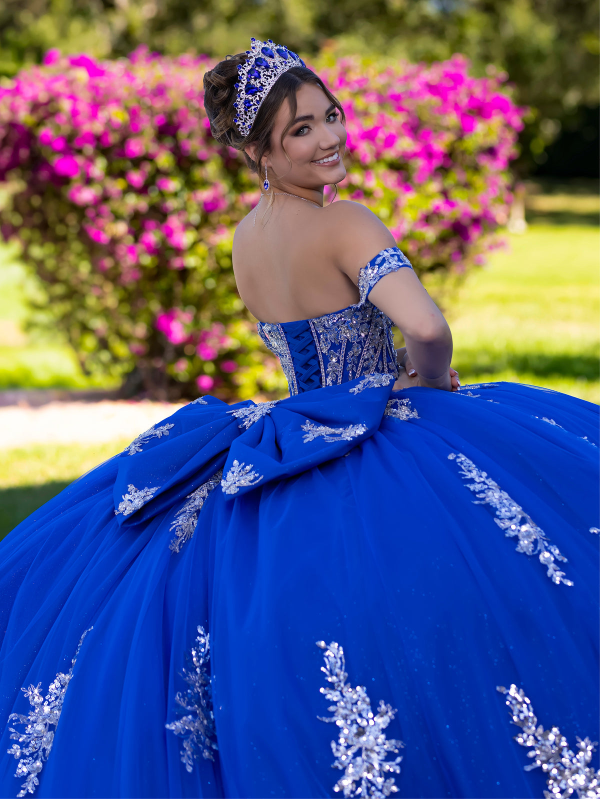 This ball gown features a detailed embroidered bodice and off-the-shoulder straps. The full, dramatic skirt is adorned with shimmering floral patterns and finishes with a large bow and flowing train.