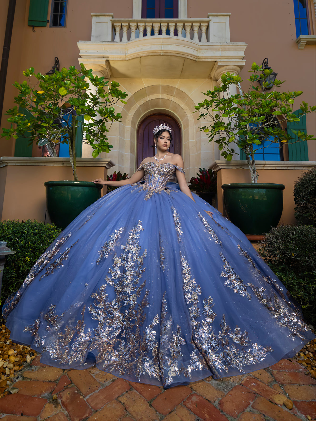 This ball gown features a detailed embroidered bodice and off-the-shoulder straps. The full, dramatic skirt is adorned with shimmering floral patterns and finishes with a large bow and flowing train.