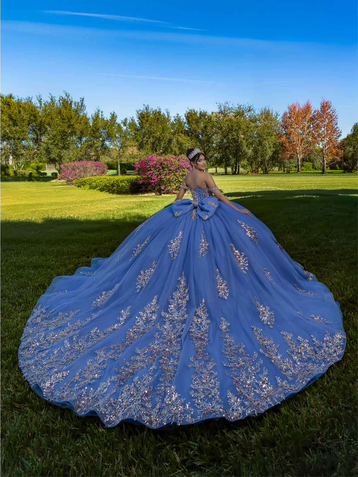 This ball gown features a detailed embroidered bodice and off-the-shoulder straps. The full, dramatic skirt is adorned with shimmering floral patterns and finishes with a large bow and flowing train.