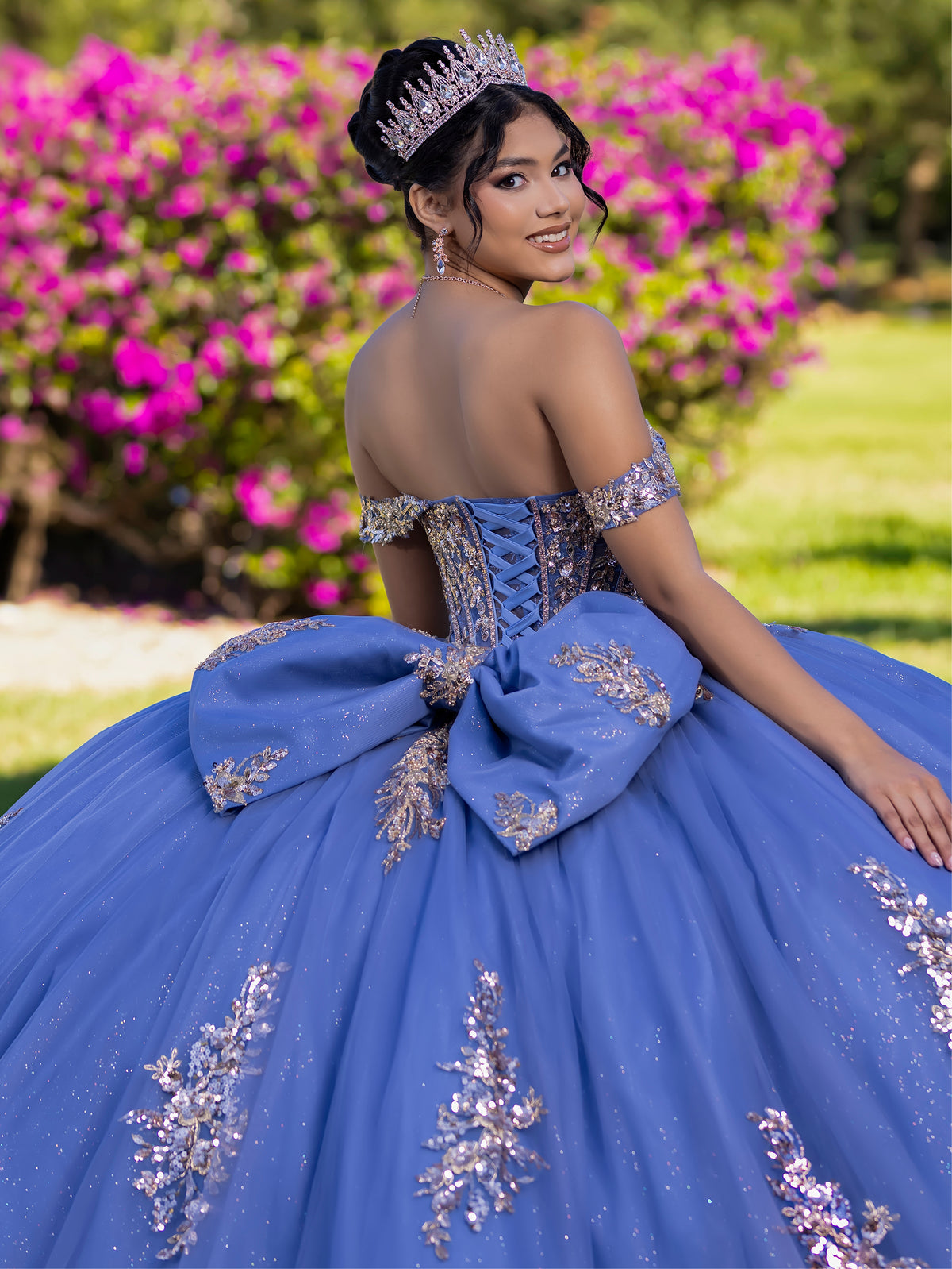 This ball gown features a detailed embroidered bodice and off-the-shoulder straps. The full, dramatic skirt is adorned with shimmering floral patterns and finishes with a large bow and flowing train.