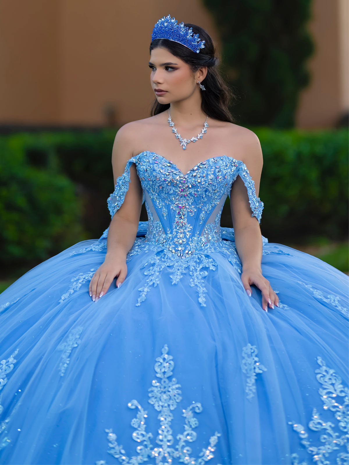 This ball gown features a richly embroidered bodice with off-the-shoulder straps. The full, voluminous skirt is decorated with shimmering floral detailing and flows into an elegant train accented with a bow at the back.