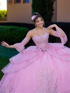 This ball gown features a fitted, embroidered bodice with off-the-shoulder sleeves. The full layered skirt includes cascading floral details and is finished with a dramatic bow and long, flowing train.