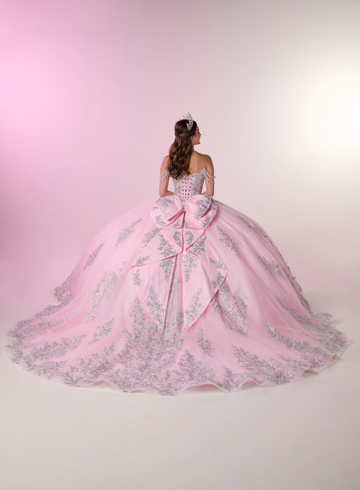 A woman viewed from behind wearing an elaborate light pink ball gown with silver floral embroidery and a large bow at the back, standing against a soft pink studio backdrop. / Enchanting ball gown with sparkling embroidery, an off-shoulder neckline, and a voluminous skirt finished with a dramatic oversized bow at the back.