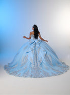 A woman viewed from behind wearing a voluminous light blue ball gown with silver floral embroidery and a large bow at the back, posed against a white studio backdrop with blue side lighting. / Enchanting ball gown with sparkling embroidery, an off-shoulder neckline, and a voluminous skirt finished with a dramatic oversized bow at the back.