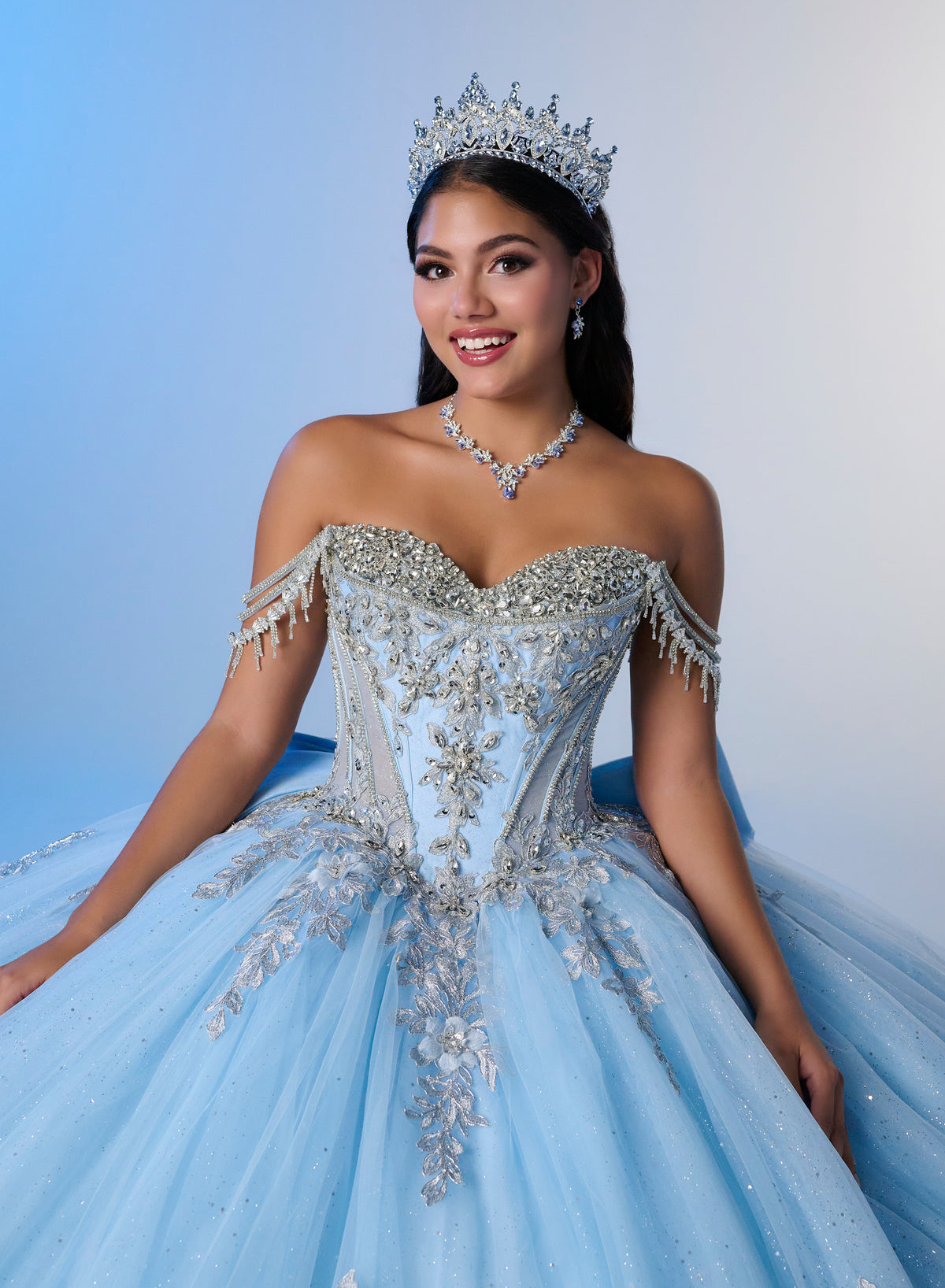 A woman wearing an elaborate off-the-shoulder light blue ball gown with silver beading and embroidery, a matching tiara, and a silver necklace, posed against a soft blue studio background. / Enchanting ball gown with sparkling embroidery, an off-shoulder neckline, and a voluminous skirt finished with a dramatic oversized bow at the back.