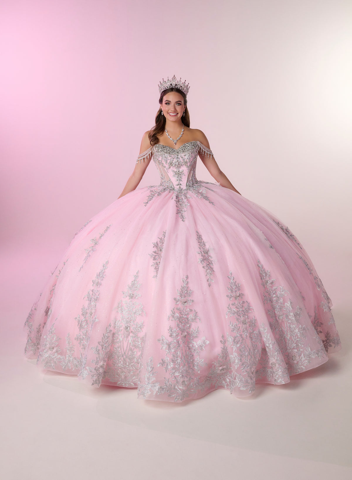 A young woman wearing a voluminous light pink ball gown with silver floral embroidery and a silver tiara, posing against a soft pink studio backdrop. / Enchanting ball gown with sparkling embroidery, an off-shoulder neckline, and a voluminous skirt finished with a dramatic oversized bow at the back.