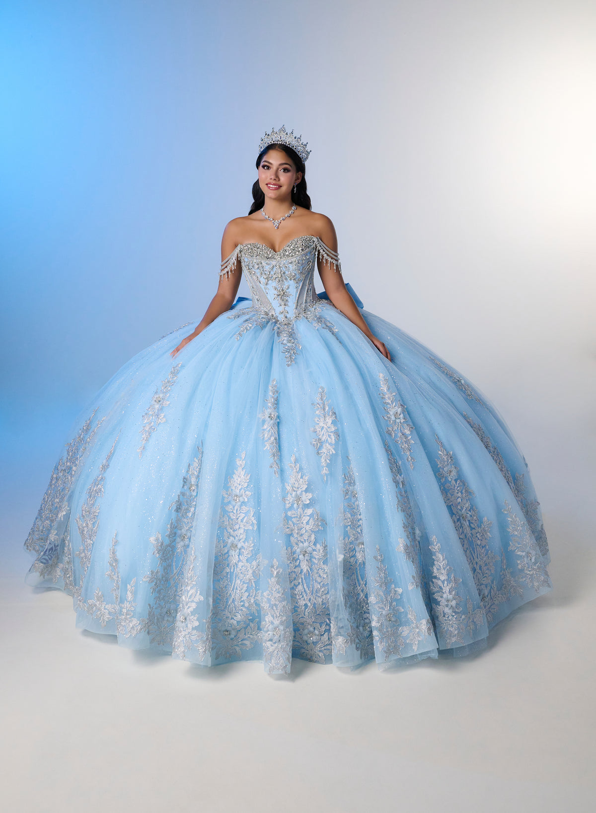 A woman wearing a voluminous light blue ball gown with silver floral embroidery and a tiara, posed against a blue-to-white studio backdrop. / Enchanting ball gown with sparkling embroidery, an off-shoulder neckline, and a voluminous skirt finished with a dramatic oversized bow at the back.