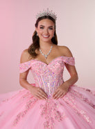 A smiling young woman wearing a sparkling off-the-shoulder pink ball gown with sequins and tulle, a silver tiara, and a matching silver necklace, posed against a soft pink studio background. / Stunning off-shoulder ball gown with shimmering embellished appliqués, a structured corset bodice, a full tulle skirt, and a dramatic oversized bow with a sweeping train.